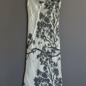 May include: A sleeveless floor-length dress in a pale sage green color featuring a dark charcoal gray tree branch silhouette print. The garment has a draped cowl neckline and a straight-cut skirt, hanging from a wooden clothes hanger.