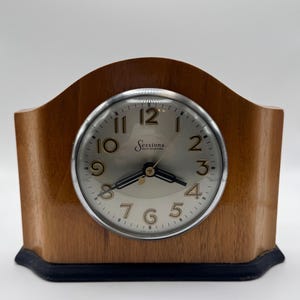 May include: A mantle clock with a light brown wooden arched case and a dark base. The circular face features a silver background, black numerals from 1 to 12, black hands, and the text Sessions Self Starting printed in the center of the dial.