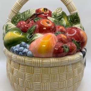 May include: A ceramic cookie jar shaped like a woven basket with a handle, featuring a lid piled high with sculpted, glossy fruits including red apples, strawberries, cherries, a green pear, and a cluster of blue grapes, all with green leaves.
