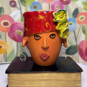 May include: A terracotta-colored ceramic head planter featuring a red patterned headpiece with yellow floral accents. The face has defined features, including pink lips and dark eyes. Silver-toned dangling hoop earrings are attached to the ears.