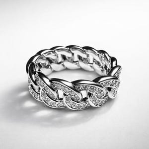 May include: A silver ring with a chain link design. The ring is encrusted with small, clear gemstones. The ring is set against a white background, highlighting its reflective surface and intricate details. A piece of jewelry.