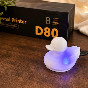 Può includere: Una luce notturna bianca a forma di anatra con una luce blu, collegata a un cavo a strisce bianche e nere. Una scatola nera dietro di essa recita "Thermal Printer Portable Tattoo Printer D80". La luce è su una superficie di legno.
