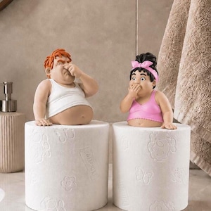 Könnte beinhalten: Zwei dekorative Figuren auf Toilettenpapierrollen. Eine Figur hat orangefarbenes Haar, trägt ein weißes Unterhemd und hält sich die Nase zu. Die andere Figur hat dunkles Haar mit einem rosa Stirnband, trägt ein rosa Oberteil und hält sich den Mund.