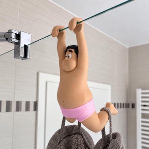 Könnte beinhalten: Eine Cartoon-Figur mit gebräunter Haut, schwarzen Haaren und rosa Unterwäsche, die an einer Glasduschwand hängt. Die Figur hält sich mit beiden Armen an der Oberkante der Glasscheibe fest, während ihre Füße ein graues, strukturiertes Handtuch stützen.