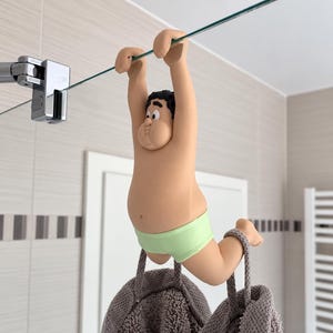 Könnte beinhalten: Eine Cartoon-Figur mit dunklem Haar, die eine hellgrüne Unterhose trägt und mit den Händen an einer Glasduschwand hängt. Die Figur hat eine rundliche Statur und ist über einem grauen, strukturierten Handtuch mit zwei Aufhängeschlaufen positioniert.