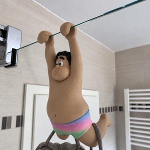 Könnte beinhalten: Eine Tonfigur eines Zeichentrickcharakters mit gebräunter Haut und schwarzen Haaren, der eine regenbogenfarbene Badehose trägt. Die Figur hängt mit den Händen an einem dünnen, horizontalen Glasregal, die Beine sind nach hinten angewinkelt.