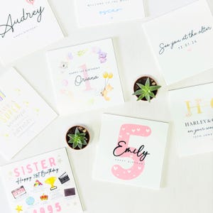 May include: A collection of white greeting cards with various designs and text, including birthday wishes and names like Audrey and Emily. Some cards feature illustrations of animals and balloons. Small potted succulents are also present.