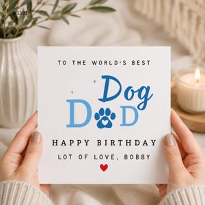 May include: A square white greeting card with black and blue text that reads TO THE WORLD'S BEST Dog Dad HAPPY BIRTHDAY LOT OF LOVE, BOBBY. The design features a blue paw print with a small white heart inside and a small red heart at the bottom.