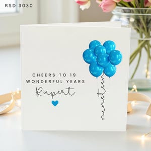 May include: A white square greeting card with the text "CHEERS TO 19 WONDERFUL YEARS", the name "Rupert", a blue heart, and a cluster of bright blue balloons. The word "nineteen" is written vertically in black script below the balloons. Code RSD 3030.