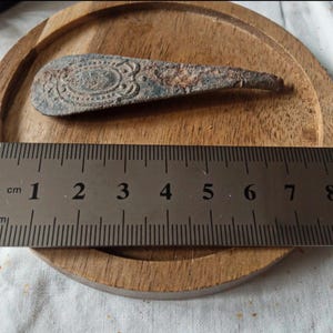 Puede incluir: Un objeto metálico antiguo y desgastado con un diseño ovalado y ornamentado en un extremo. El objeto mide aproximadamente 7,6 cm de largo y termina en punta. Descansa sobre una superficie de madera, con una regla para la escala.