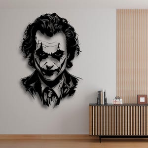 Könnte beinhalten: Ein schwarz-weißes Metall-Wandkunstwerk, das den Joker darstellt. Das Kunstwerk zeigt das ikonische Make-up und die Frisur der Figur. Der Joker trägt einen Anzug und eine Krawatte. Die Kunst ist an einer hellgrauen Wand angebracht.