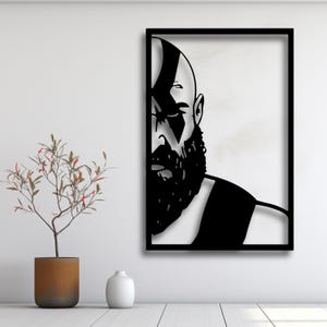 Op de afbeelding: Zwart metalen wandkunst met een gestileerd portret van een bebaarde man met een opvallende markering op zijn hoofd. Het kunstwerk is omlijst in een zwarte rechthoekige lijst, die contrasteert met de witte achtergrond. Een modern woondecoratie-item.