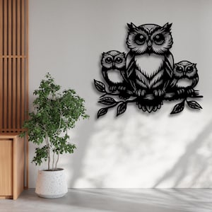 Op de afbeelding: Zwart metalen wandkunstwerk met drie uilen op een tak met bladeren. De middelste uil is groter, geflankeerd door twee kleinere uilen. Het kunstwerk is gemonteerd op een lichtgekleurde muur, met een plant in een pot op de voorgrond.