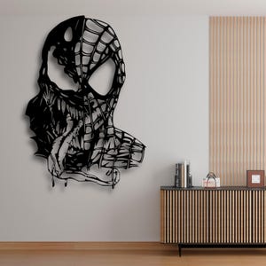 May include: Black metal wall art depicting a split face design, half Spider-Man and half Venom. The artwork features intricate web patterns and sharp, angular details, creating a striking contrast. The piece is mounted on a neutral wall.