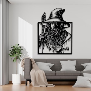 Könnte beinhalten: Schwarze Metall-Wandkunst, die einen Zauberer mit langem Bart, Stab und Hut darstellt. Das Kunstwerk ist in einem schwarzen Quadrat gerahmt und an einer weißen Wand befestigt. Der Zauberer hält eine Pfeife. Im Vordergrund befinden sich ein Sofa und eine Pflanze.