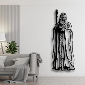 May include: Black metal wall art depicting a robed figure with a staff. The artwork features a long beard and a staff with a fork-like top. The figure is standing, and the design is a silhouette.