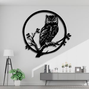 Könnte beinhalten: Schwarze Metall-Eulen-Wandkunst. Die Eule sitzt auf einem Ast in einem kreisförmigen Rahmen. Das Kunstwerk ist eine Silhouette mit komplizierten Details der Federn der Eule und der Äste. Die Wandkunst ist an einer weißen Wand angebracht.