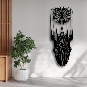 May include: Black metal wall art featuring an abstract design with intricate cut-out details. The artwork has a symmetrical shape with a crown-like top and a pointed bottom. It is mounted on a white wall, adding a modern touch to the space.