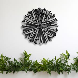 May include: A black metal spider web wall decoration with a spider in the center. The web is a circular design with radiating lines. The background is a white wall with green plants at the bottom.