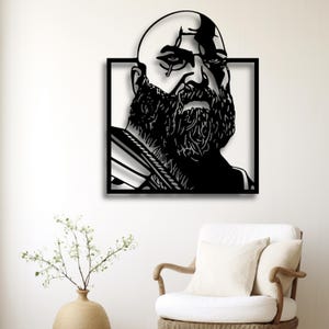 Op de afbeelding: Zwarte metalen wandkunst met een kale man met een volle baard en een intense uitdrukking. Het kunstwerk is omlijst in een zwart vierkant. Een opvallend, modern decoratiestuk voor in huis.