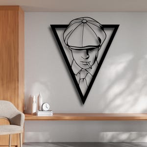 Könnte beinhalten: Schwarze Metall-Wandkunst, die einen Mann mit flacher Kappe und Krawatte zeigt, der eine Zigarette raucht, innerhalb eines schwarzen Dreiecksrahmens. Das Kunstwerk ist an einer hellgrauen Wand angebracht, darunter ein Holzregal und dekorative Gegenstände.