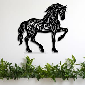 May include: Black metal horse wall art with intricate floral design. The horse is depicted in a dynamic pose, with one leg raised. The artwork is mounted on a white wall, with green plants at the bottom.