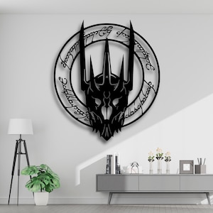 Puede incluir: Una decoración de pared de metal negro que representa una cara estilizada con una corona y una inscripción circular. La obra de arte está montada en una pared blanca. Una lámpara y una planta en maceta están en primer plano, con un gabinete gris y decoración en el fondo.