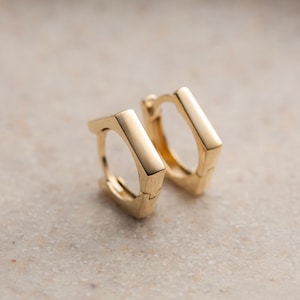 May include: A pair of gold-colored, geometric hoop earrings. Each earring has a rectangular shape with rounded edges, creating a modern and minimalist design. The earrings are displayed on a neutral, textured surface.
