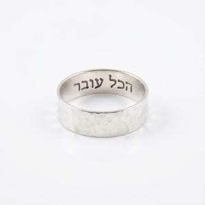 May include: A silver ring with a hammered texture and the Hebrew inscription "This Too Shall Pass" engraved on the outer band. The ring is set against a white background.