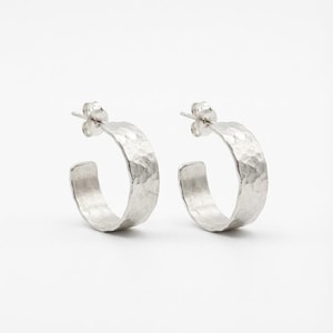 Pode incluir: Um par de brincos de argola prateados com textura martelada. Os brincos têm fecho de pino e um design espesso e curvo. A superfície texturizada capta a luz. Um acessório de joalharia clássico.