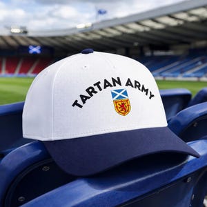 May include: White baseball cap with a navy blue brim. The cap features the text "TARTAN ARMY" in black, along with the Scottish flag and a shield emblem. The cap is sitting on a blue stadium seat.