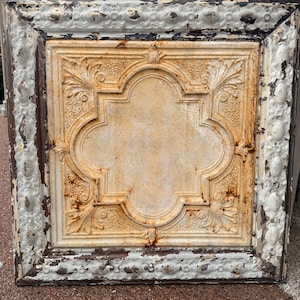 Puede incluir: Una baldosa de techo cuadrada de metal antiguo con una apariencia desgastada y envejecida. El diseño central presenta una forma de cuatro hojas con detalles de relieve floral ornamentado. El marco es blanco con pintura descascarillada, revelando metal oscuro debajo y una pátina oxidada y envejecida.