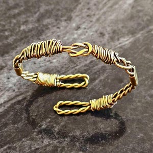 May include: A handcrafted gold-toned bracelet with a unique design. The bracelet features a twisted wire pattern and a hook closure. The open cuff design allows for easy wear. The jewelry is displayed on a dark, textured surface.