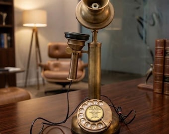 May include: A brass candlestick telephone with a circular rotary dial base. The center of the dial features text reading TO CALL, RECEIVE, JAPAN TOP, HEADSET SOUND, FULL DIAL, TO CALL, AND LET GO. A black cord connects the handset to the base.