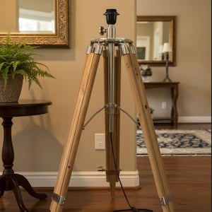 Puede incluir: Una lámpara de pie con trípode de madera y portalámparas negro. La lámpara tiene tres patas de madera de color claro con detalles metálicos plateados. La lámpara está sobre un suelo de madera noble.