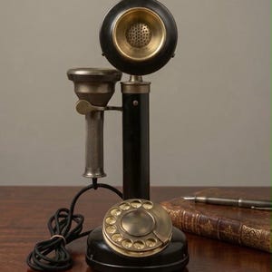 May include: A vintage-style candlestick telephone featuring a black cylindrical body, a brass rotary dial base, and a matching brass-accented receiver. The device includes a black coiled cord and a metallic handset cradle mounted on the central pillar.