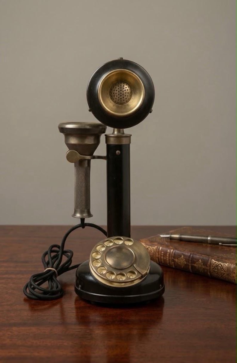 May include: A vintage-style candlestick telephone featuring a black cylindrical body, a brass rotary dial base, and a matching brass-accented receiver. The device includes a black coiled cord and a metallic handset cradle mounted on the central pillar.