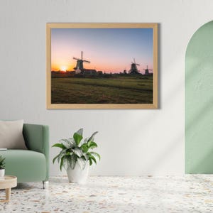 May include: A framed print of a sunset over a field with several windmills. The sky transitions from orange to blue. The windmills are silhouetted against the setting sun, and the field is green.