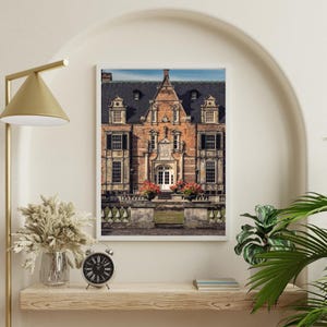 Puede incluir: Una impresión enmarcada de un imponente edificio de ladrillo con un tejado oscuro y múltiples ventanas. El edificio tiene una puerta blanca y está flanqueado por jardineras. La impresión está en una estantería de madera con un reloj y un jarrón.