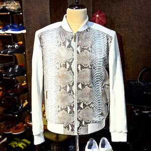 Op de afbeelding: Een witte bomberjack met slangenpatroon en ritssluiting wordt op een mannequin gepresenteerd. Witte sneakers staan op een houten standaard voor de jas. De jas heeft witte mouwen en afwerking.