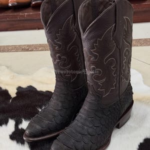 Puede incluir: Un par de botas de vaquero marrones oscuras con costuras intrincadas y un patrón texturizado en forma de escamas en la parte inferior. Las botas tienen una punta puntiaguda y un tacón apilado, sobre una alfombra de piel de vaca en blanco y negro.