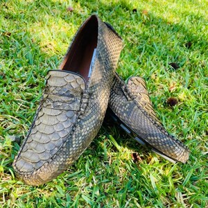 Puede incluir: Un par de mocasines texturizados de color verde oliva con un patrón escamoso. Los zapatos están hechos de un material que se asemeja a la piel de serpiente. Los mocasines están sobre hierba verde.