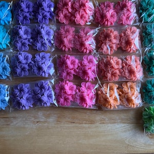 Pode incluir: Uma variedade de decorações coloridas em forma de flor em vários tons de azul, roxo, rosa, laranja e verde, embaladas em plástico transparente. As flores estão dispostas sobre uma superfície de madeira.