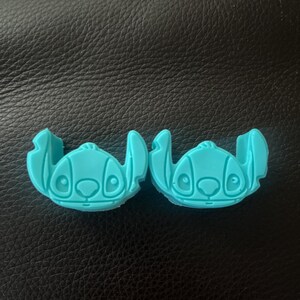 Può includere: Due oggetti blu chiaro a forma di Stitch. Ognuno ha un viso dettagliato con grandi occhi, un naso piccolo e una bocca larga. Gli oggetti hanno grandi orecchie e sono probabilmente oggetti decorativi o funzionali.