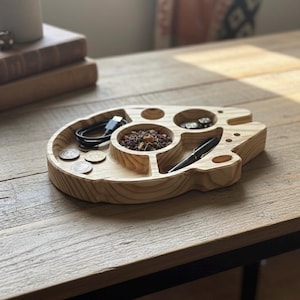 May include: A handmade wooden tray in a light wood finish, shaped like a spaceship. It features multiple compartments holding a USB cable, coins, a pen, and a small bowl filled with dried herbs. The tray sits on a wooden table.