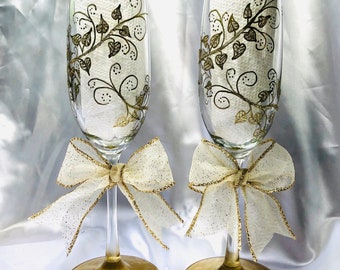 May include: Two champagne flutes with gold leaf painted floral designs and gold glitter accents. Each flute is tied with a white and gold bow.