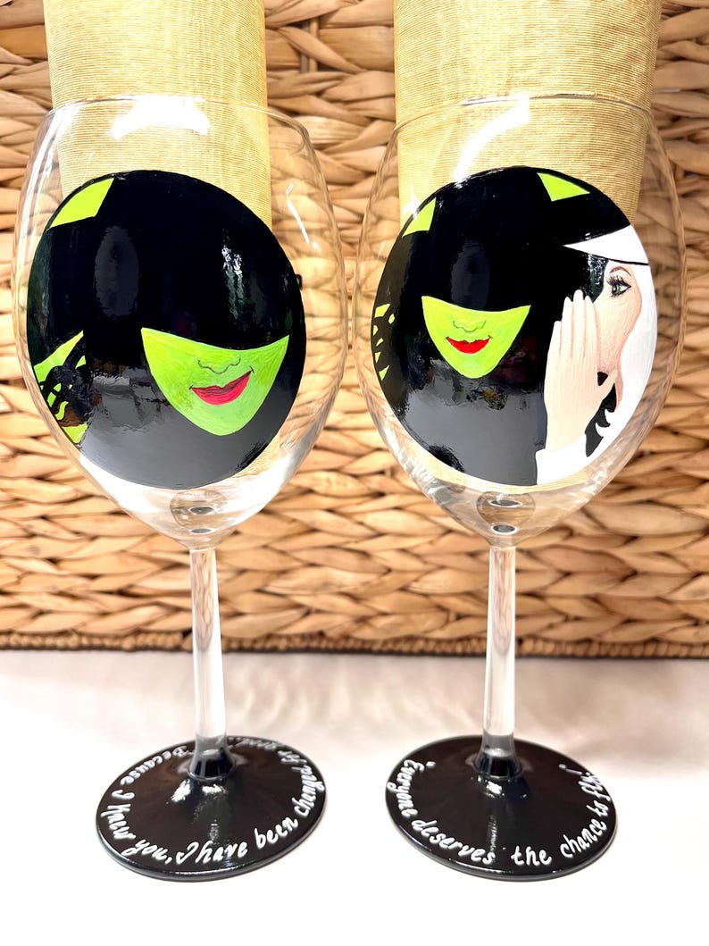 May include: Two black and white wine glasses with green and red painted designs. The glasses have black stems and bases with white text. The text on the left glass reads "I knew you, I have been changed". The text on the right glass reads "Everyone deserves the chance to fly".