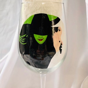 May include: A hand-painted wine glass with a black and green image of the Wicked Witch of the West from the Wizard of Oz.