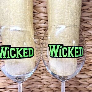 May include: Two clear glass wine glasses with black and green "Wicked" decals on the front. The glasses are sitting on a woven wicker surface.