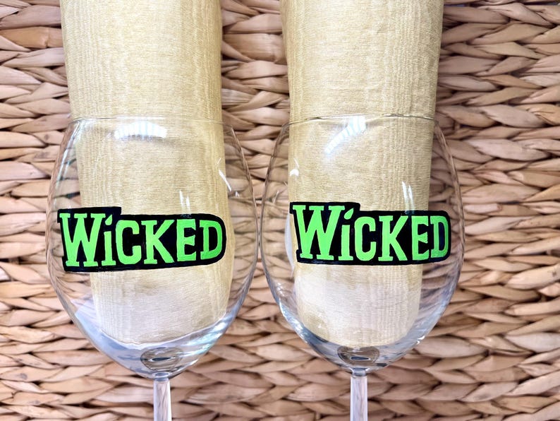 May include: Two clear glass wine glasses with black and green "Wicked" decals on the front. The glasses are sitting on a woven wicker surface.